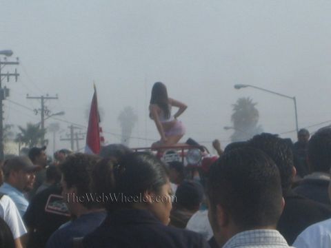 1990 Ford Bronco - dense crowd gathers in foggy morning light around a red off-road vehicle on which a woman in shorts is perched.