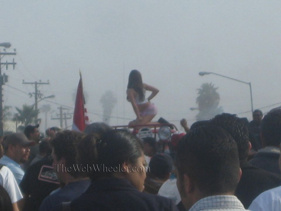 Crowd at outdoor event with woman in shorts sitting atop a red vehicle in foggy haze