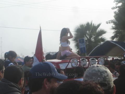 1990 Ford Bronco - woman sits atop the front of a red off-road race truck above a crowd, with a flag and a Baja 1000 banner in the foggy background.