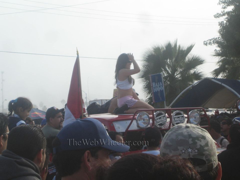 Woman seated on hood of red off-road truck above a crowd, flag and Baja 1000 sign nearby
