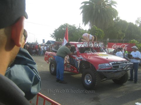 1990 Ford Bronco - red off-road race truck is parked curbside at an event with a Coca-Cola banner; a man in green leans against it as spectators watch.