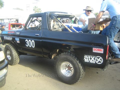 1990 Ford Bronco - black lifted Ford Bronco race truck marked 300 with BFGoodrich and ATX Series decals sits in a pit area as crew sort supplies.