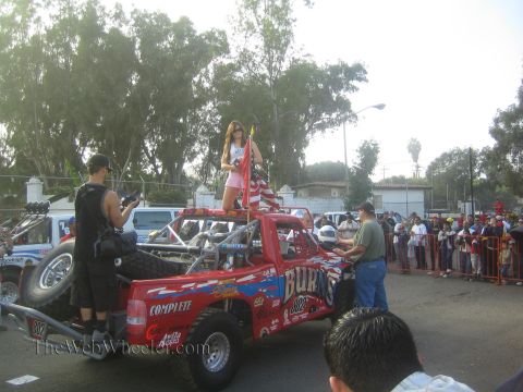 1990 Ford Bronco - woman stands in the bed of a red off-road race truck holding a flag while a photographer films and spectators line a barricade.