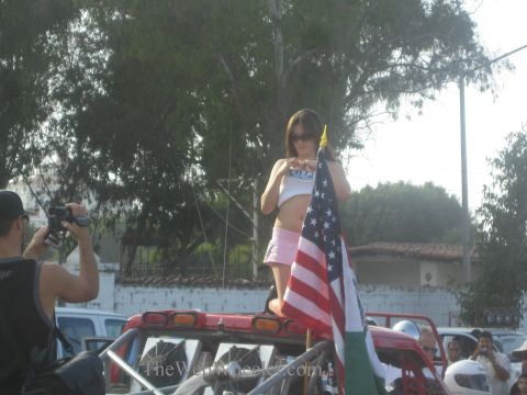 1990 Ford Bronco - woman in a white top stands on a vehicle holding a US flag as a man photographs her against a backdrop of trees and parked cars.