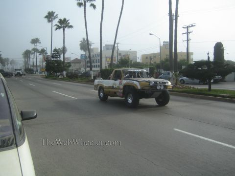1990 Ford Bronco - white and gold off-road prerunner truck drives down a foggy palm-lined boulevard past buildings and parked cars.