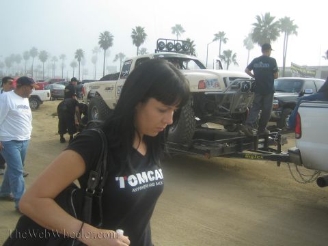 1990 Ford Bronco - woman in a black Tomcat T-shirt walks past a white off-road race truck strapped to a Big Tex trailer in a sandy lot under palms.