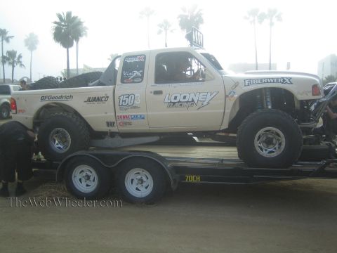 1990 Ford Bronco - tan Looney Racing off-road truck with FiberwerX and BFGoodrich graphics sits strapped to a black trailer in a palm-lined lot.