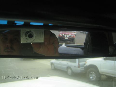 1990 Ford Bronco - man photographs himself in a vehicle's rearview mirror with a silver compact camera, a red truck and white SUV reflected behind.
