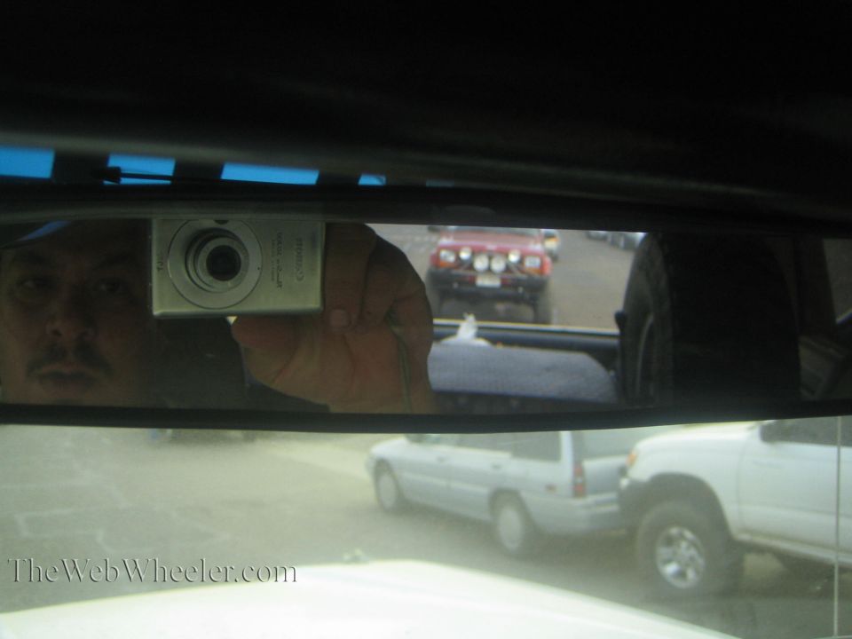 Rearview mirror selfie of a man holding a silver camera, red truck visible behind