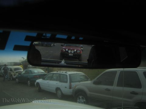 1990 Ford Bronco - rearview mirror frames a red off-road truck while a row of parked cars and a white SUV fill the foreground through the windshield.