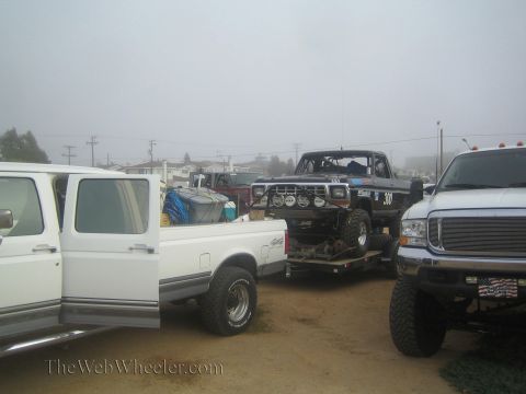 1990 Ford Bronco - black Ford race truck numbered 300 sits on a trailer between two white pickups in a foggy lot, one a lifted Super Duty.