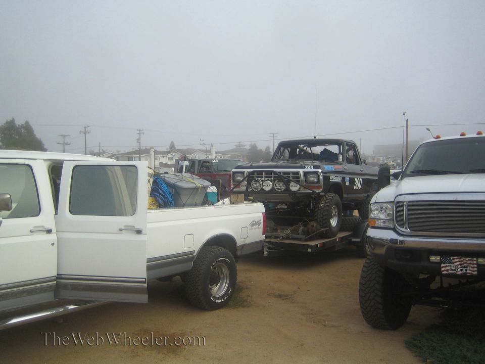 Black Ford race truck number 300 on a trailer between two white pickups in fog