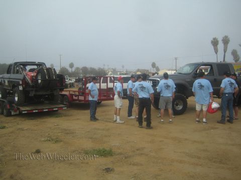 1990 Ford Bronco - group of men in matching light-blue team shirts stand in a foggy dirt lot beside off-road trucks, one loaded on a trailer.
