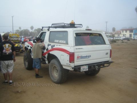 1990 Ford Bronco - white Ford Bronco lettered Score Medical with red graphics and a roof rack sits in a foggy dirt staging lot as a man stands beside it.