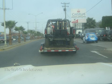 1990 Ford Bronco - Seen through a windshield, an off-road truck carrying spare tires rides a flatbed trailer down a foggy street past a Cazuelas sign.