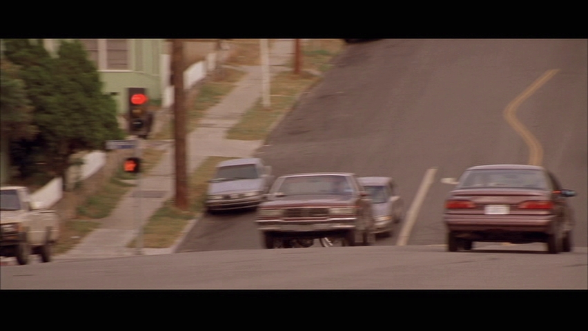 Cars descending a steep residential street in a film still, two sedans mid-chase