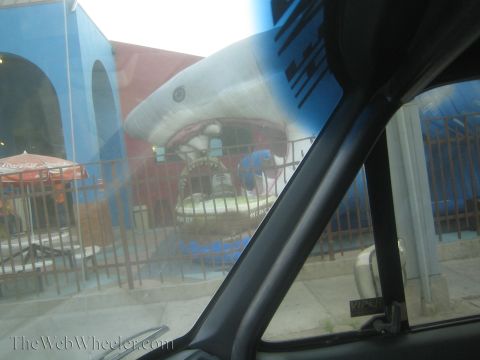 1990 Ford Bronco - Through a car window a large shark sculpture juts from a blue building facade above a fenced patio with Coca-Cola umbrellas.