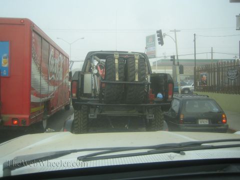 1990 Ford Bronco - Through a windshield, a black off-road truck loaded with spare tires on a trailer sits in traffic beside a red Coca-Cola delivery truck.