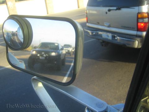 1990 Ford Bronco - vehicle's side mirror reflects a lifted black off-road truck in a line of traffic, with a silver SUV passing alongside.