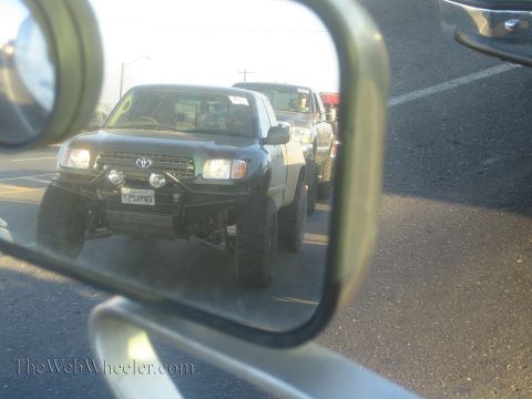 1990 Ford Bronco - side mirror reflects a lifted black Toyota pickup with a front brush guard and prerunner lights waiting in a line of traffic.