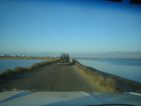 1990 Ford Bronco - off-road truck towed on a trailer travels a narrow dirt causeway flanked by calm blue water with distant mountains and buildings.