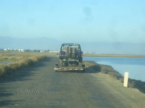 1990 Ford Bronco - lifted off-road truck loaded on a flatbed trailer is towed along a dirt levee road beside still water at dusk.