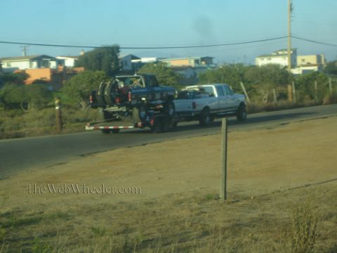 1990 Ford Bronco - white pickup tows a flatbed trailer carrying a blue lifted off-road truck along a dirt shoulder near houses.
