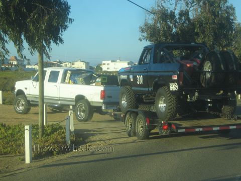 1990 Ford Bronco - white Ford pickup is parked beside a tandem-axle trailer loaded with a black lifted off-road race truck numbered 31.