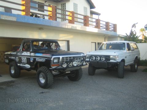 1990 Ford Bronco - black number 300 Ford Bronco off-road race truck and a white lifted Ford Bronco sit parked on pavement outside a house.
