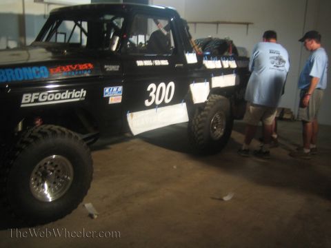 1990 Ford Bronco - black BFGoodrich-sponsored number 300 off-road race truck sits in a garage as two men stand inspecting it.