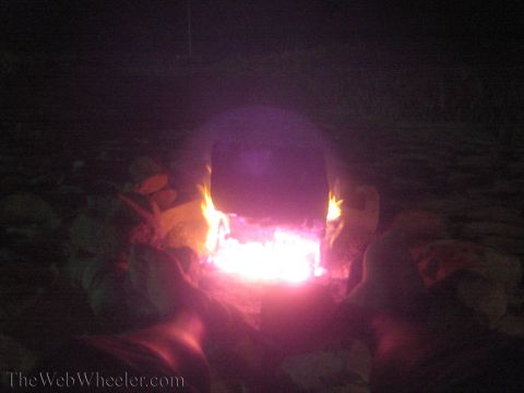 1990 Ford Bronco - glowing campfire burns with pink and orange flames on a dark beach at night, a dim figure behind it.