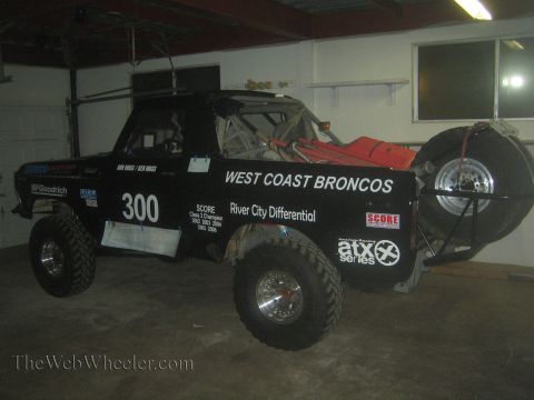 1990 Ford Bronco - black West Coast Broncos number 300 off-road race truck with a spare tire sits parked inside a garage.