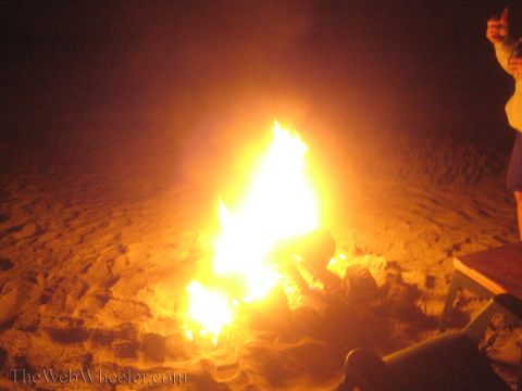 1990 Ford Bronco - bonfire burns on a sandy beach at night while a person stands at the right with an arm raised.