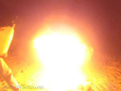 1990 Ford Bronco - beach fire flares into a bright white-orange burst at night as a person in light clothing stands at the left.