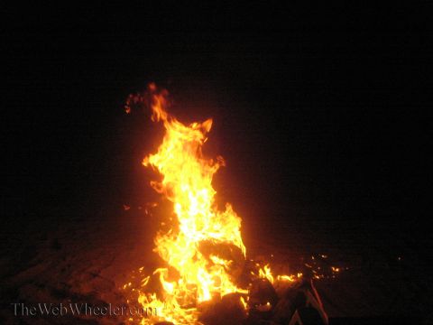 1990 Ford Bronco - tall orange-and-yellow campfire flame rises from glowing embers against a black night background.