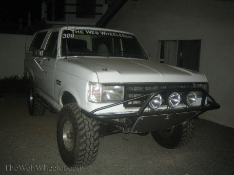 1990 Ford Bronco - white lifted Ford Bronco with a number 300 windshield banner, front light bar and round driving lights sits outside at night.