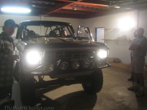 1990 Ford Bronco - lifted off-road truck with headlights and driving lights blazing sits in a garage as two men stand nearby.