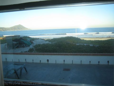 1990 Ford Bronco - Window view overlooking a sandy beach, green dune vegetation and ocean waves with a headland and rising sun on the horizon.