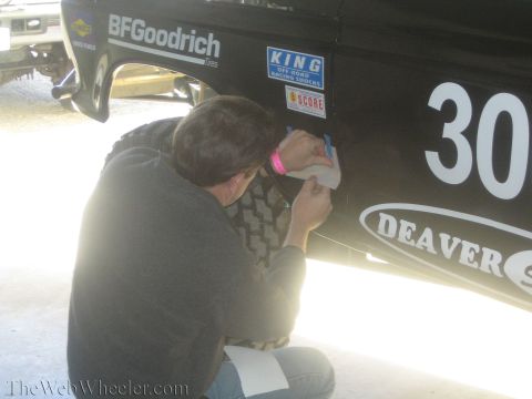 1990 Ford Bronco - man kneels to place a decal on the black flank of a race vehicle marked 300 with BFGoodrich, KING and Deaver sponsor stickers.