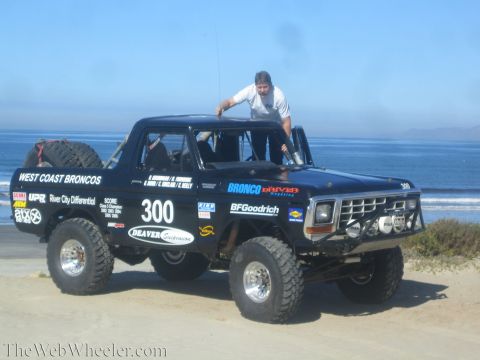 1990 Ford Bronco - man stands in the roll-caged cockpit of black race truck 300 lettered West Coast Broncos, parked on sand beside the ocean.