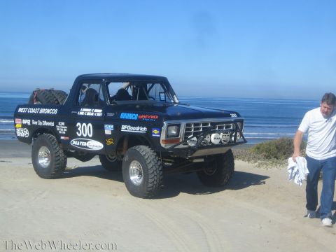 1990 Ford Bronco - Black off-road race truck 300 with West Coast Broncos livery sits on the sand by the surf as a man in blue walks past at right.