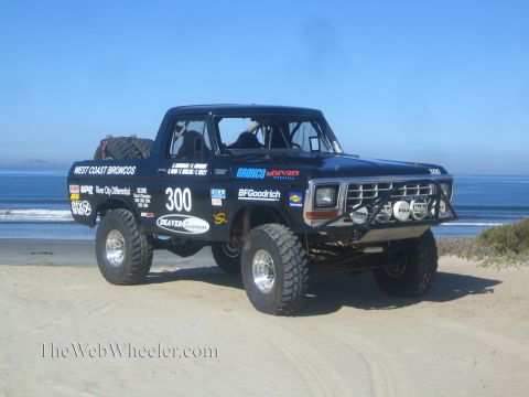 1990 Ford Bronco - black number 300 off-road race truck with West Coast Broncos and BFGoodrich livery sits on a sandy beach beside calm ocean.