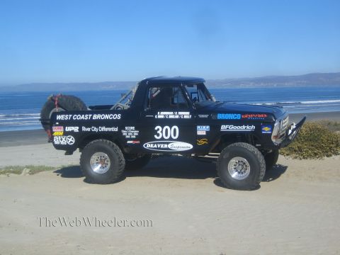 1990 Ford Bronco - Profile of black race truck 300 with West Coast Broncos livery and rear spare tire, parked on the beach with mountains behind.