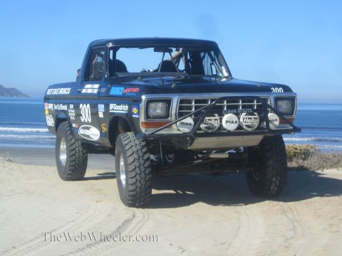 1990 Ford Bronco - Front three-quarter view of black race truck 300 fitted with a Hella and PIAA light bar, sitting on sand beside the ocean.