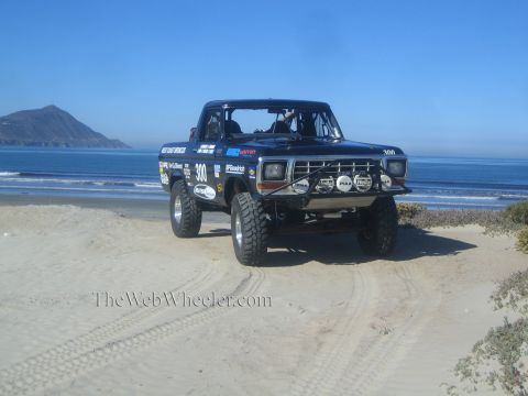 1990 Ford Bronco - black number 300 off-road race truck sits on the sand near tire tracks, with ocean surf and a rocky headland behind it.