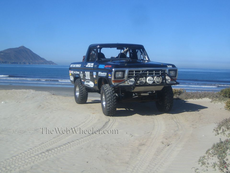 Black number 300 off-road race truck parked on a beach with a headland in the background