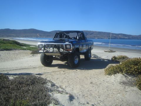 1990 Ford Bronco - lifted dark off-road truck with a front light bar sits on a beach dune amid iceplant, with a long shoreline and hills beyond.