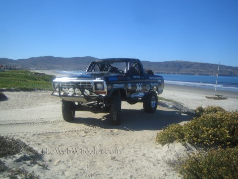 1990 Ford Bronco - dark number 300 race truck with auxiliary light pods sits on a sandy dune with iceplant, ocean and coastal hills behind.