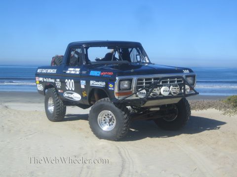1990 Ford Bronco - Front three-quarter view of a black number 300 off-road race truck with PIAA and Hella lights, parked on a beach by the ocean.