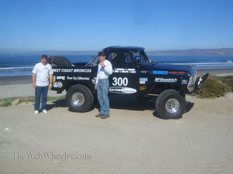1990 Ford Bronco - Two men in white shirts pose beside the black number 300 West Coast Broncos race truck on the sand with the ocean behind.